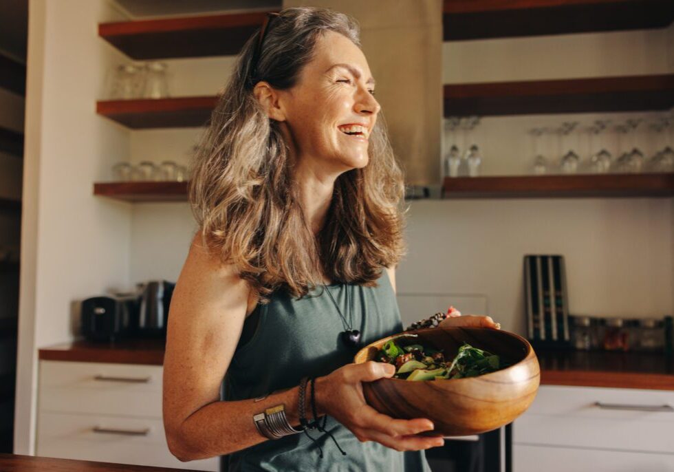 Aging woman smiling happily while holding a buddha bowl in her kitchen. Happy senior woman serving herself a healthy vegan meal at home. Mature woman taking care of her body with a plant-based diet.